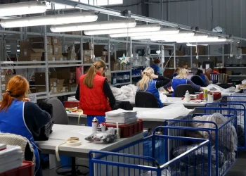 Trabalhadoras em fábrica de vestuário organizam e embalam peças de roupa em ambiente industrial iluminado, com prateleiras cheias de caixas ao fundo.