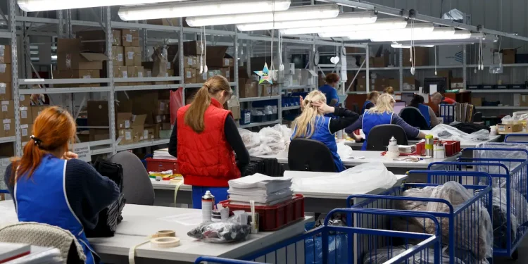 Trabalhadoras em fábrica de vestuário organizam e embalam peças de roupa em ambiente industrial iluminado, com prateleiras cheias de caixas ao fundo.