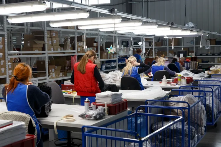 Trabalhadoras em fábrica de vestuário organizam e embalam peças de roupa em ambiente industrial iluminado, com prateleiras cheias de caixas ao fundo.