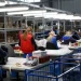 Trabalhadoras em fábrica de vestuário organizam e embalam peças de roupa em ambiente industrial iluminado, com prateleiras cheias de caixas ao fundo.